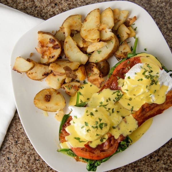 Eggs Benedict with bacon, spinach, and tomato, served with roasted potato wedges on a white plate.